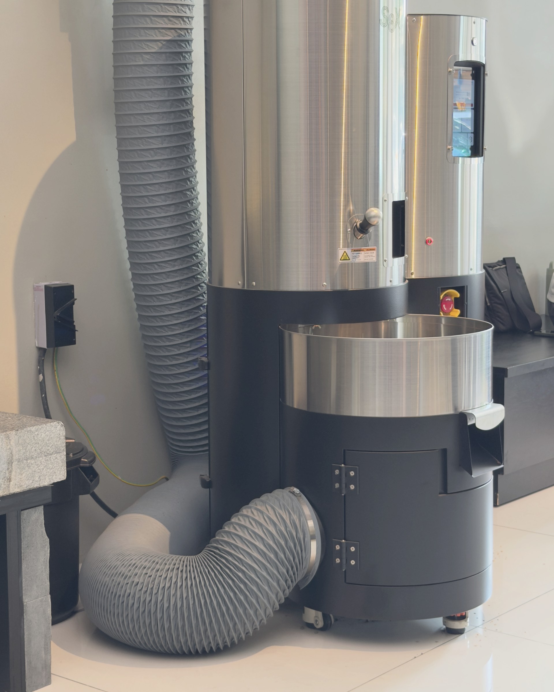 A small-batch drum roaster with its extraction ducting, in a white room